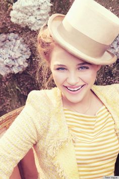 a young woman wearing a hat and smiling for the camera with flowers in the background