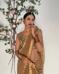 a woman in a brown sari and gold jewelry standing next to a tree with pink flowers