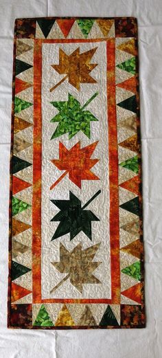 a quilted wall hanging with leaves on it's sides and an orange strip at the bottom