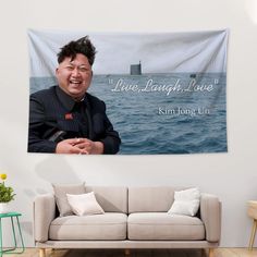 a man is smiling while sitting on a couch in front of a wall tapestry with the words live laugh love