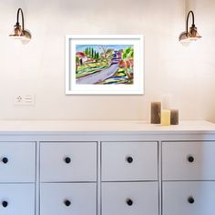 a painting hangs on the wall above a white dresser with drawers and two lamps over it