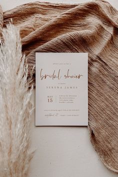 a white and gold wedding card on top of a brown blanket next to a feather