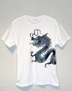 The Live Like a Dragon Graphic T-shirt is a regular fit classic crew neck, crafted from organic white cotton fabric for a crisp feel. Featuring Rob Pepper's hand drawn Dragon this elegant t-shirt is hand made and so there may be some slight variations in size and positioning of the graphic. The lead time for this t-shirt is 7 days. Unisex Material:  100% certified organic ringspun combed cotton. Ribbed collar. Taped neck. Side seams. Twin needle stitching. Crew neck Hand-drawn graphics  Weight: White Organic Cotton T-shirt With Graphic Design, Graphic Tee Cotton T-shirt With Custom Artwork, Cotton T-shirt With Custom Artwork And Relaxed Fit, Cotton T-shirt With Custom Artwork In Relaxed Fit, White Cotton T-shirt With Custom Artwork, White Organic Cotton Crew Neck Shirt, Artistic White T-shirt With Screen Print, White Cotton Tops With Custom Artwork, White Organic Cotton T-shirt