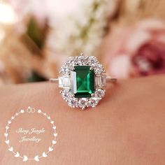 an emerald and diamond ring sitting on top of a woman's arm with flowers in the background