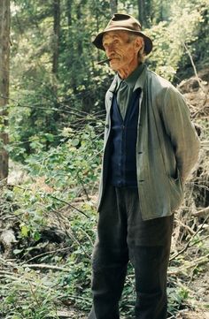 an old man standing in the middle of a forest wearing a hat and jacket with his hands on his hips