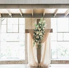 a cross decorated with flowers and greenery in front of large windows at an indoor wedding venue