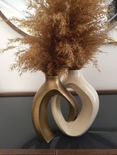 a white vase with some brown flowers in it on a table next to a mirror