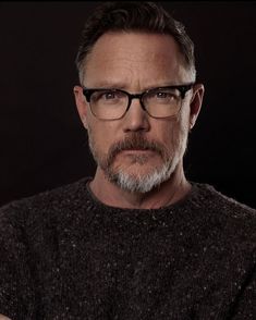 a man wearing glasses and a sweater looks at the camera with an intense look on his face