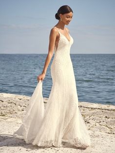 a woman in a white dress is walking on the sand near the water and holding a bag