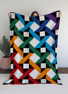 a woman holding up a colorful quilt on top of a white table next to a potted plant