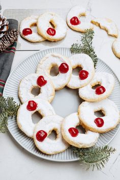 several cookies with cherries are arranged on a plate next to pine cones and evergreen branches