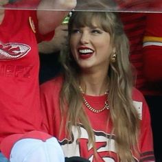 a woman with long hair wearing a red shirt and blue jeans is laughing while sitting in the stands at a football game