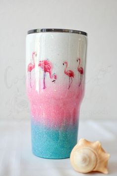 a pink and blue cup with flamingos painted on it next to a seashell