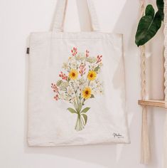 a white tote bag with sunflowers and other flowers on it next to a plant