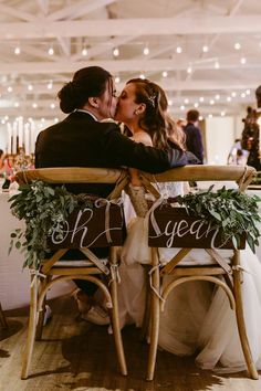 a bride and groom kiss on their wedding day at the reception table with chairs decorated with greenery