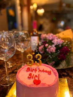 a pink birthday cake sitting on top of a table next to wine glasses and flowers