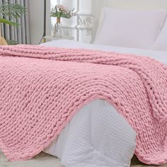 a pink blanket is on top of a bed with white sheets and pillows, next to a potted plant