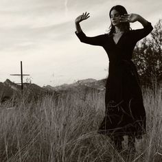 a woman in a long black dress is standing in tall grass with her hands up