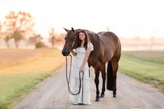 a woman standing next to a horse on a dirt road in front of a field