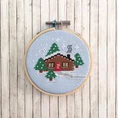 a cross - stitch christmas scene with a house and trees in the snow on a wooden background