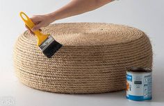 a person using a paint brush on top of a jute ottoman with a can of paint next to it