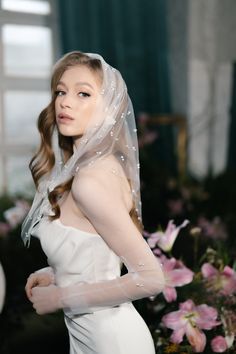 a woman in a white wedding dress and veil with flowers behind her head looking off to the side