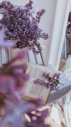 an open book sitting on top of a wooden chair next to purple flowers in vases