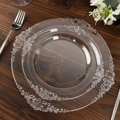 an empty glass plate sitting on top of a wooden table next to silverware and flowers