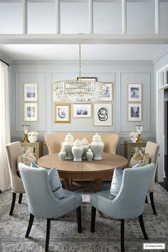 a dining room table with blue chairs and pictures on the wall above it, in front of a chandelier