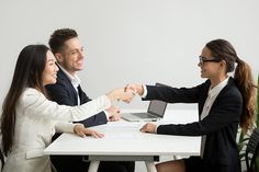 three business people shaking hands in front of a laptop computer on a table with another person sitting at the desk