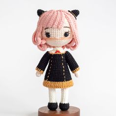 a crocheted doll with pink hair and black dress standing on a wooden base