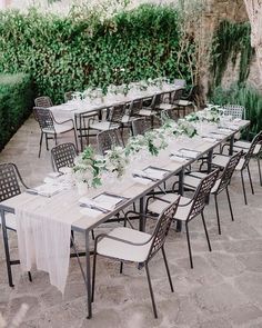 an outdoor dining table set up with white linens and green centerpieces, surrounded by greenery