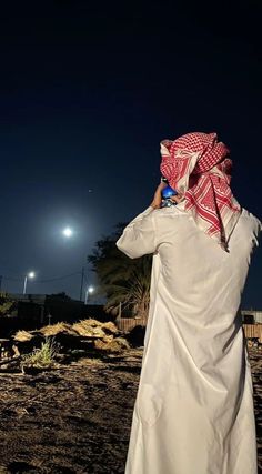 a man standing on top of a dirt field under a dark sky with a cell phone in his hand