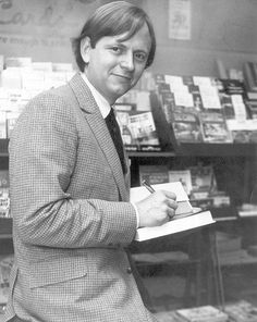 a man in a suit and tie writing on a note book at a library with stacks of books behind him