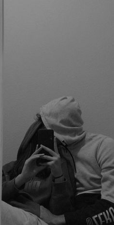 black and white photograph of person sitting on bed looking at cell phone with hood up