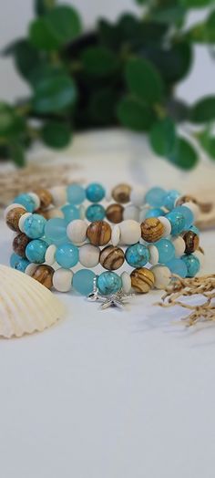 two bracelets with blue beads and shells on a table next to a plant in the background