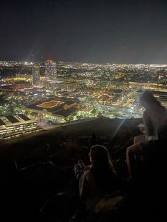 two people sitting on top of a hill looking at the city lights in the distance