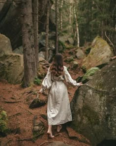 a woman in a white dress is walking through the woods with large rocks and trees