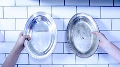 two hands holding silver dishes in front of white tiled backsplashes on a wall