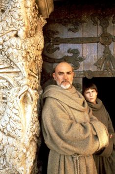 an older man standing next to a young woman in front of a carved stone wall