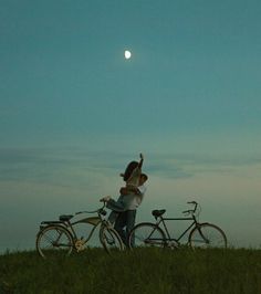 two people are standing next to their bikes in the grass at night with the moon behind them