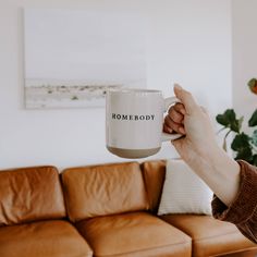 a person holding a coffee mug with the word somebody on it in front of a couch