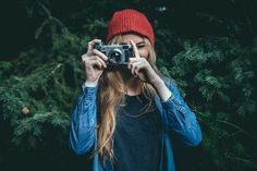 a woman taking a photo with her camera in front of some pine trees and evergreens