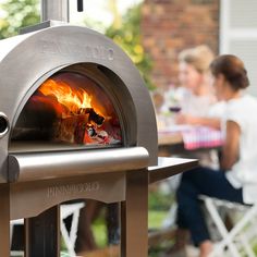 a woman sitting in front of an outdoor pizza oven with flames coming out of it