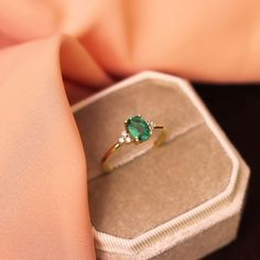 an emerald and diamond ring sitting on top of a velvet covered box, with a pink fabric behind it