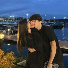 a man and woman kissing in front of the water at night with boats docked behind them