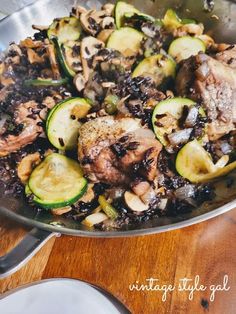 a pan filled with meat and vegetables on top of a wooden table next to silverware