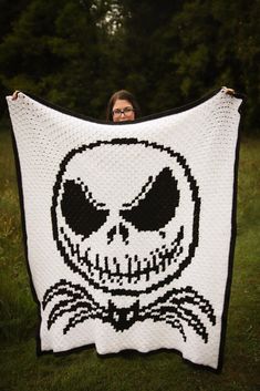 Woman holds up a large crocheted blanket of Jack Skellington. The blanket is in black and white, and features Jack's head with a smirk on his face and signature bat bow tie. The woman is standing in front of a dark, ominous looking cluster of trees. Nightmare Before Christmas Crochet, Daffodil Craft, Bat Bow, Crochet Pour Halloween, C2c Blanket, C2c Crochet Pattern Free, Yarn Weights, Yarn Project, Throw Blanket Pattern