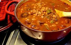 a red pot filled with beans and meat on top of a stove next to a wooden spoon