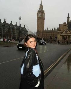 a woman standing on the side of a road in front of a building with a clock tower